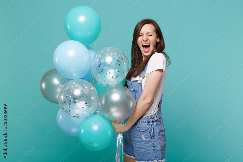 Portrait of crazy screaming funny young woman in denim clothes celebrating, holding colorful air balloons isolated on blue turquoise wall background. Birthday holiday party, people emotions concept. Portrait of crazy screaming funny young woman in denim clothes celebrating, holding colorful air balloons isolated on blue turquoise wall background. Birthday holiday party, people emotions concept.