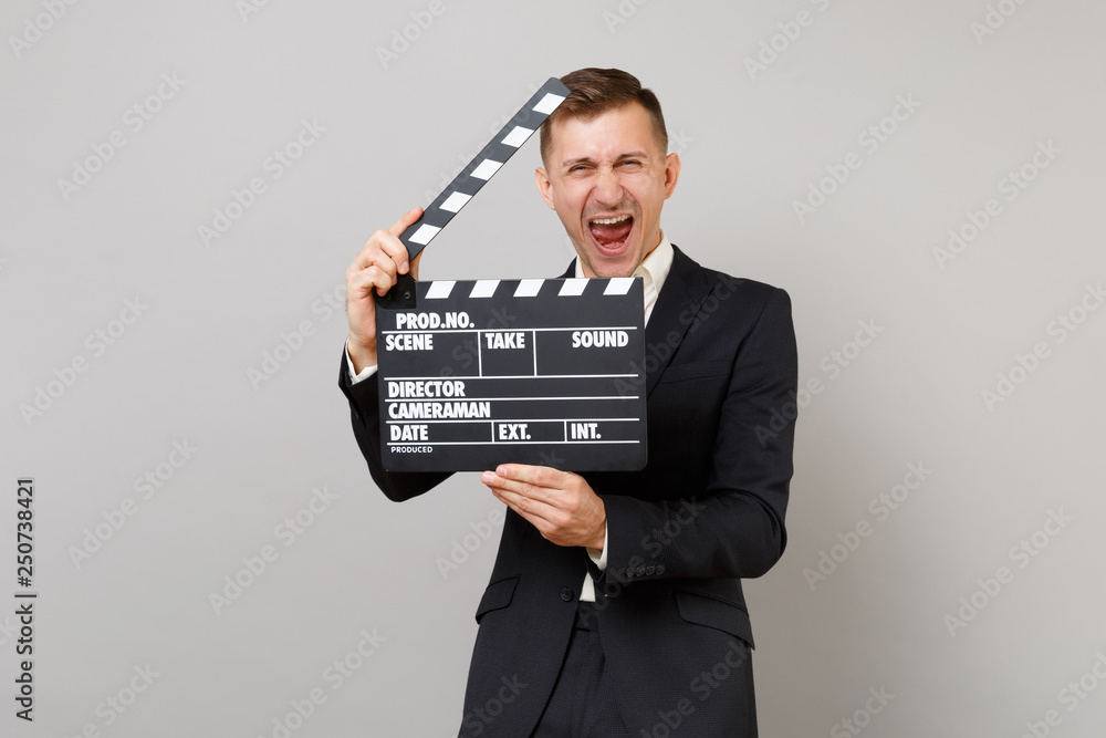 Overjoyed young business man in suit keeping mouth wide open holding classic black film making clapperboard isolated on grey background. Achievement career wealth business concept. Mock up copy space. Overjoyed young business man in suit keeping mouth wide open holding classic black film making clapperboard isolated on grey background. Achievement career wealth business concept. Mock up copy space.