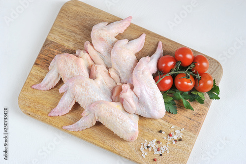 Raw chicken wings on a wooden board. The composition is complemented by cherry tomatoes, parsley and spices. White background. Close-up. View from above.