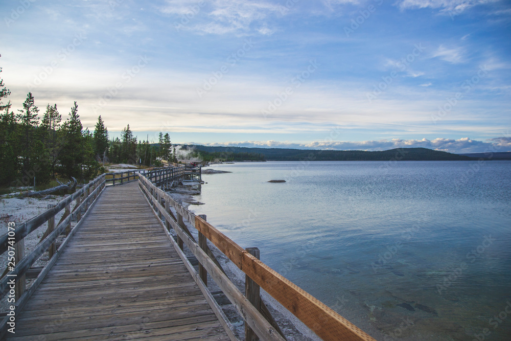 Wooden pier on the beach of Yellowstone lake. Mountains view on background. Small forest on the side. Blue sky with some clouds and blue lake with geysers. 
