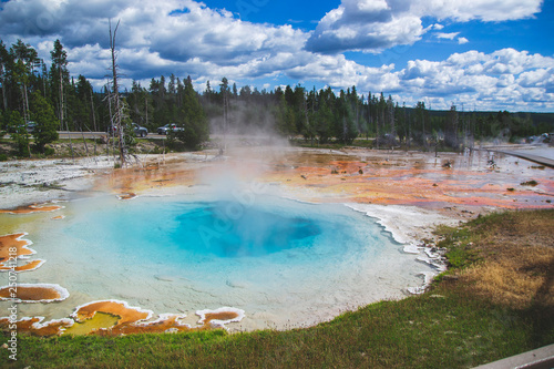Orange blue geyser in Yellowstone national park. Pool of spring in the green forest with airy clouds on blue sky. Wooden pier for walking in geysers area. National park in United States.