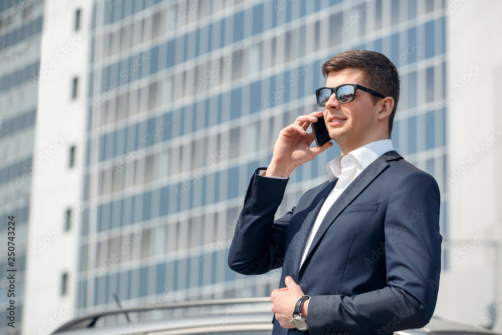 Young man in sunglasses standing near car talking on smartphone joyful Young man in sunglasses standing near car talking on smartphone joyful