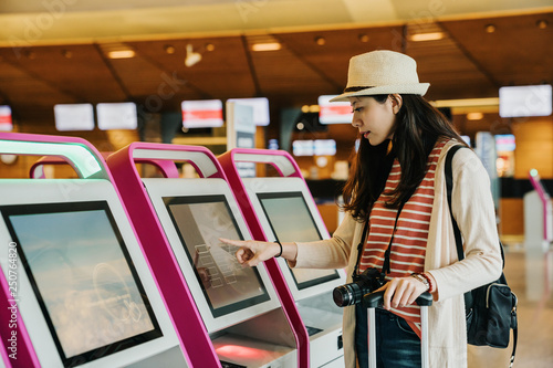 young asian female business traveler using self service check in machine at airport lobby indoor. girl photographer carrying camera finger touching screen choosing picking on auto counter concept. photo