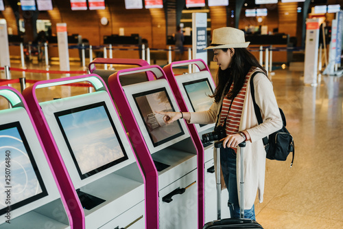 Young chinese woman in straw hat standing in international airport doing self check-in. travel lady carrying camera backpack and luggage finger clicking on auto machine indoors in lobby near counter. photo