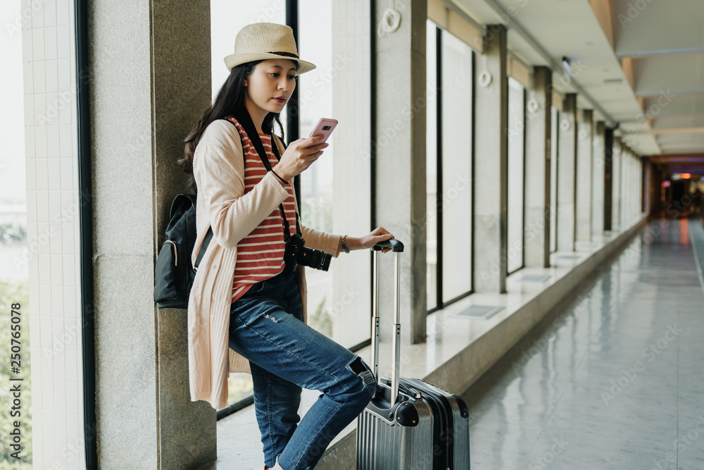 Young casual asian female traveler at airport terminal holding smartphone device looking searching online timetable schedule delay time of airplane. girl relax on wall in departure gate window. Young casual asian female traveler at airport terminal holding smartphone device looking searching online timetable schedule delay time of airplane. girl relax on wall in departure gate window.