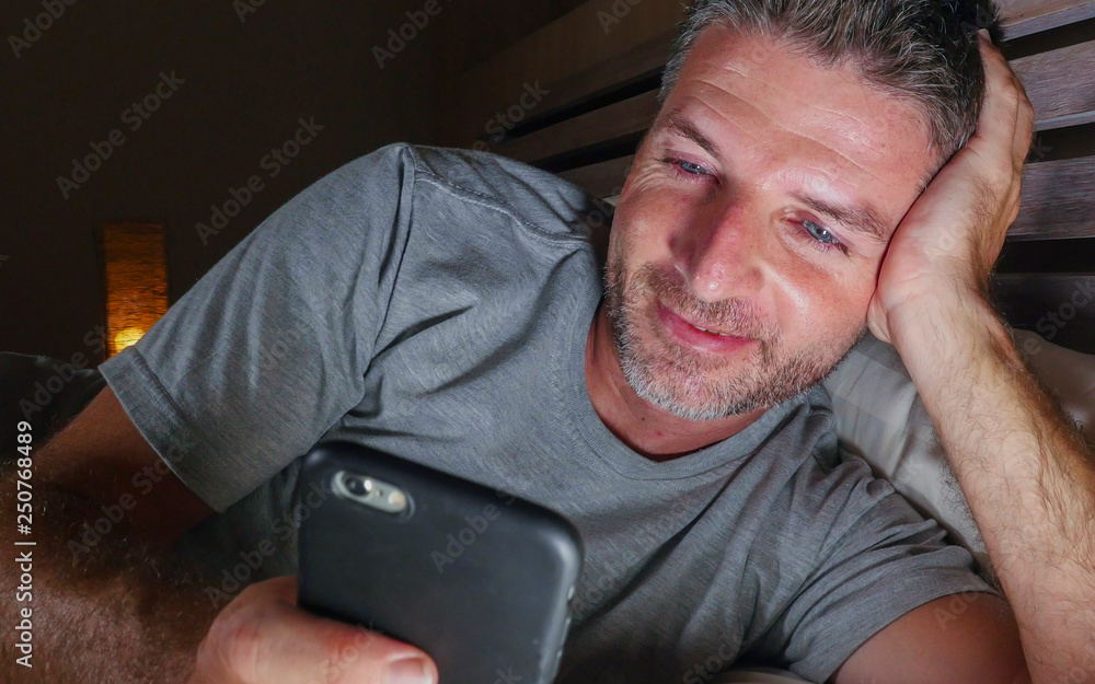 Obraz premium young handsome and attractive happy man networking with mobile phone late at night in bed smiling relaxed enjoying social media or online dating app in internet addiction