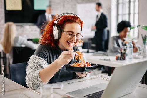 Cheerful businesswoman eating salad and listening music on headphones in the office.
