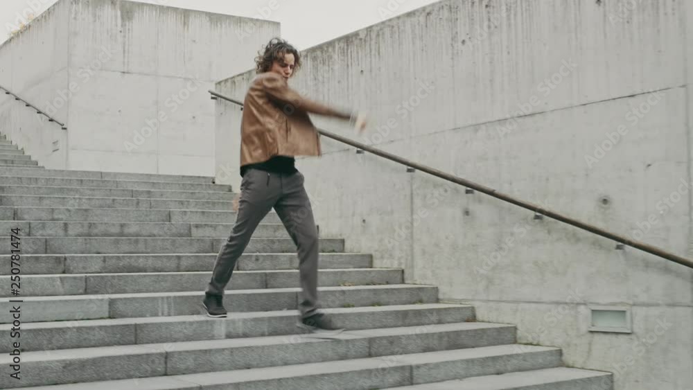 Cheerful and Happy Young Man with Long Hair Actively Dancing While Walking Down the Stairs. He's Wearing a Brown Leather Jacket. Scene Shot in an Urban Concrete Park. Day is Bright. Cheerful and Happy Young Man with Long Hair Actively Dancing While Walking Down the Stairs. He's Wearing a Brown Leather Jacket. Scene Shot in an Urban Concrete Park. Day is Bright.