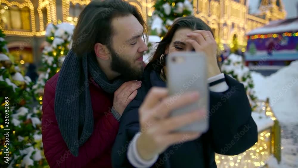 A beautiful young girl and her handsome boyfriend are photographed on a smartphone outdoors in the city Park with dozens of illuminated Christmas decorations around. Happy and healthy relationship. A beautiful young girl and her handsome boyfriend are photographed on a smartphone outdoors in the city Park with dozens of illuminated Christmas decorations around. Happy and healthy relationship.