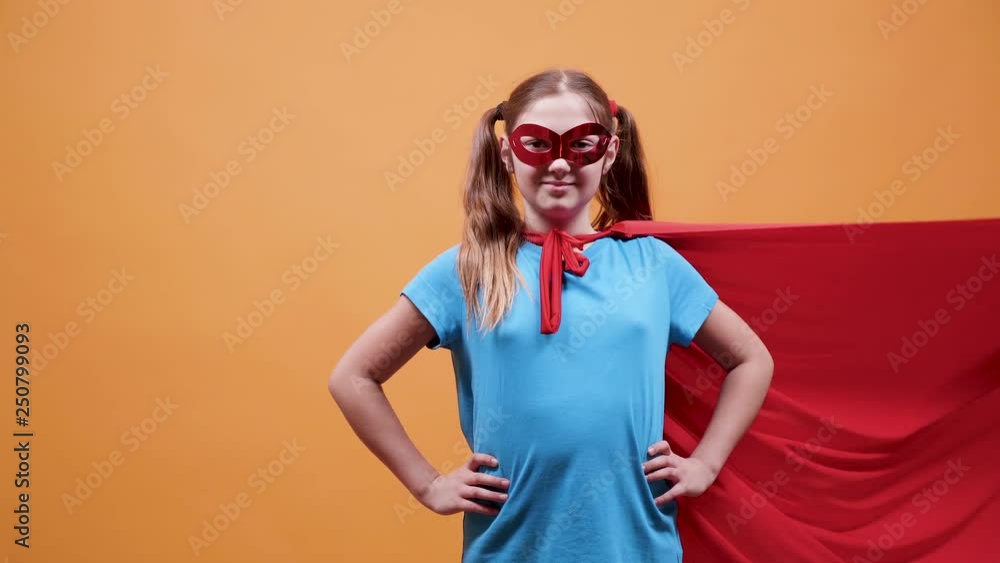 Superhero young girl with a red cloak and a mask to cover her face. Confident look at the camera. Isolated on orange background. Superhero young girl with a red cloak and a mask to cover her face. Confident look at the camera. Isolated on orange background.