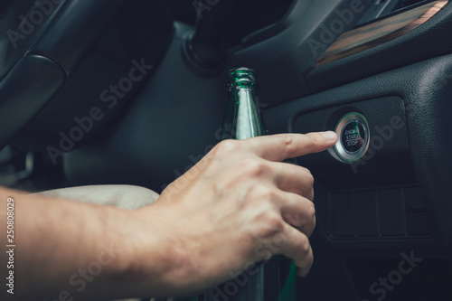 The man hand is holding a beer bottle and pressing the engine start button.