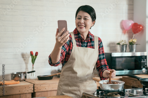 young asian housewife having video phone call on cellphone laughing chatting while cooking handmade chocolate melting in pot on stove stir by wooden spoon in decorated kitchen for valentine day. photo