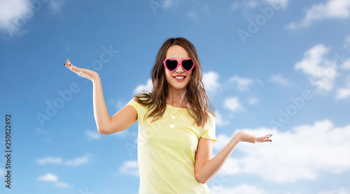 summer, valentine's day and people concept - smiling young woman or teenage girl in heart-shaped sunglasses holding something on palms over blue sky and clouds background