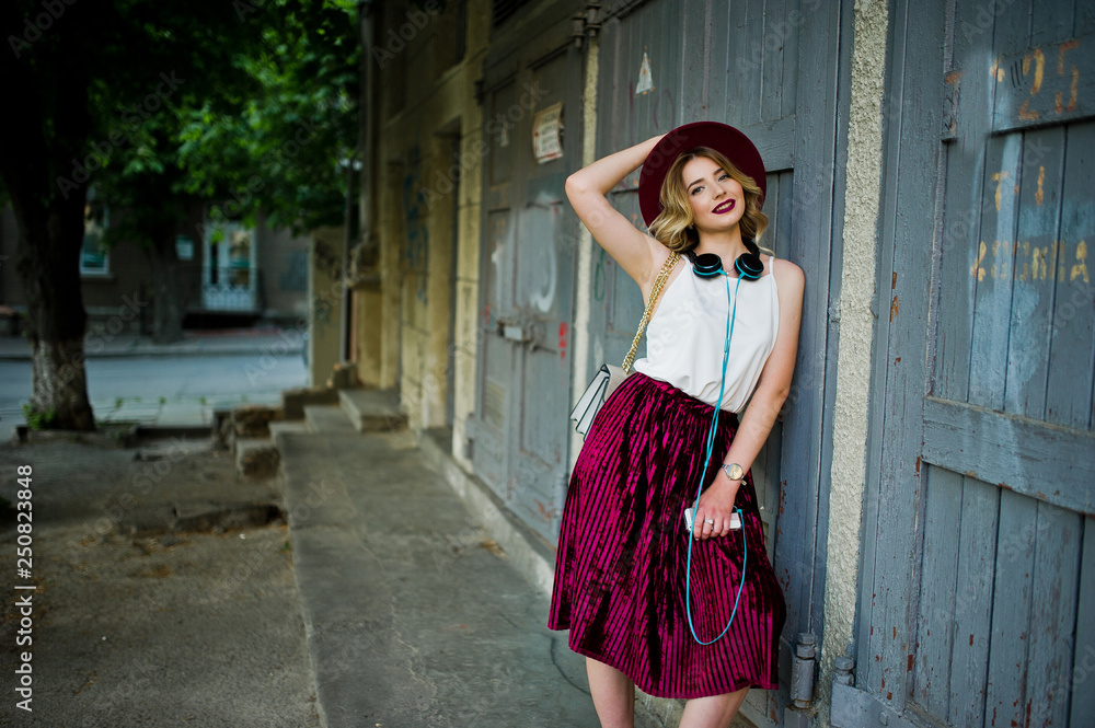 Obraz premium Fashionable and beautiful blonde model girl in stylish red velvet velour skirt, white blouse and hat, posed with phone and earphones.