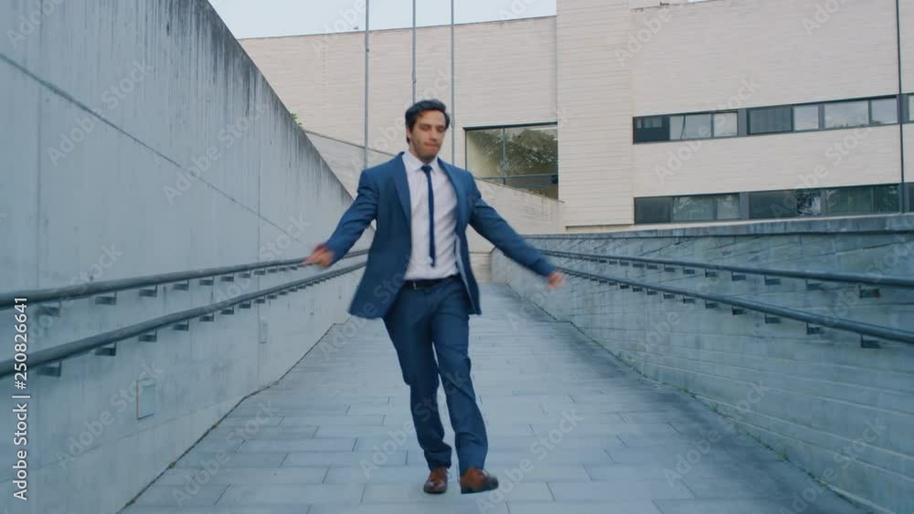 Cheerful and Happy Businessman Actively Dancing and Jumping While Walking Down a Concrete Urban Path. He's Wearing a Suit. Scene Shot Next to Business Center. Day is Bright. Cheerful and Happy Businessman Actively Dancing and Jumping While Walking Down a Concrete Urban Path. He's Wearing a Suit. Scene Shot Next to Business Center. Day is Bright.
