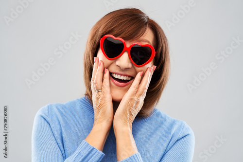 valentine's day, summer and old people concept - portrait of smiling senior woman in red heart-shaped sunglasses over grey background