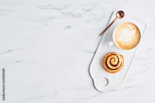 Freshly baked cinnamon roll with spices and cocoa filling and coffee or cappuccino with latte art on white serving plate over white marble background. Top view. Copy space for text. Swedish breakfast. photo