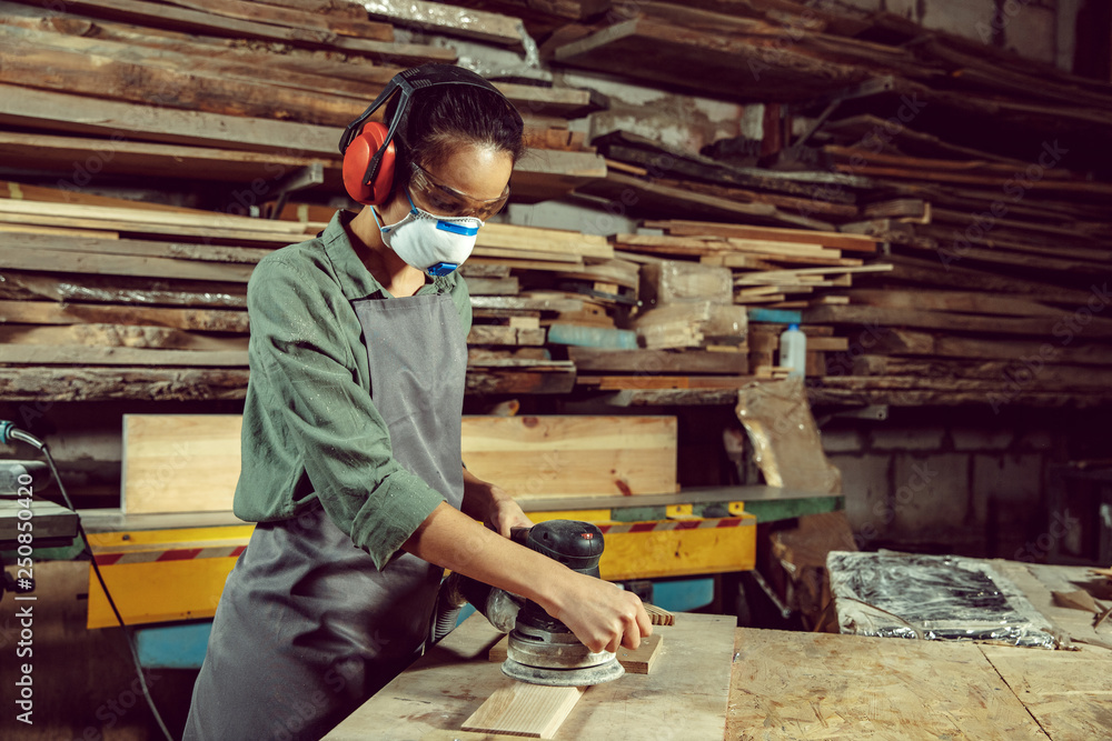 Busy and serious craftswoman grinding timbers with special machine. Beautiful woman wearing safety glasses. Concept of joiner's shop and woodworking. Gender equality. Male profession Busy and serious craftswoman grinding timbers with special machine. Beautiful woman wearing safety glasses. Concept of joiner's shop and woodworking. Gender equality. Male profession