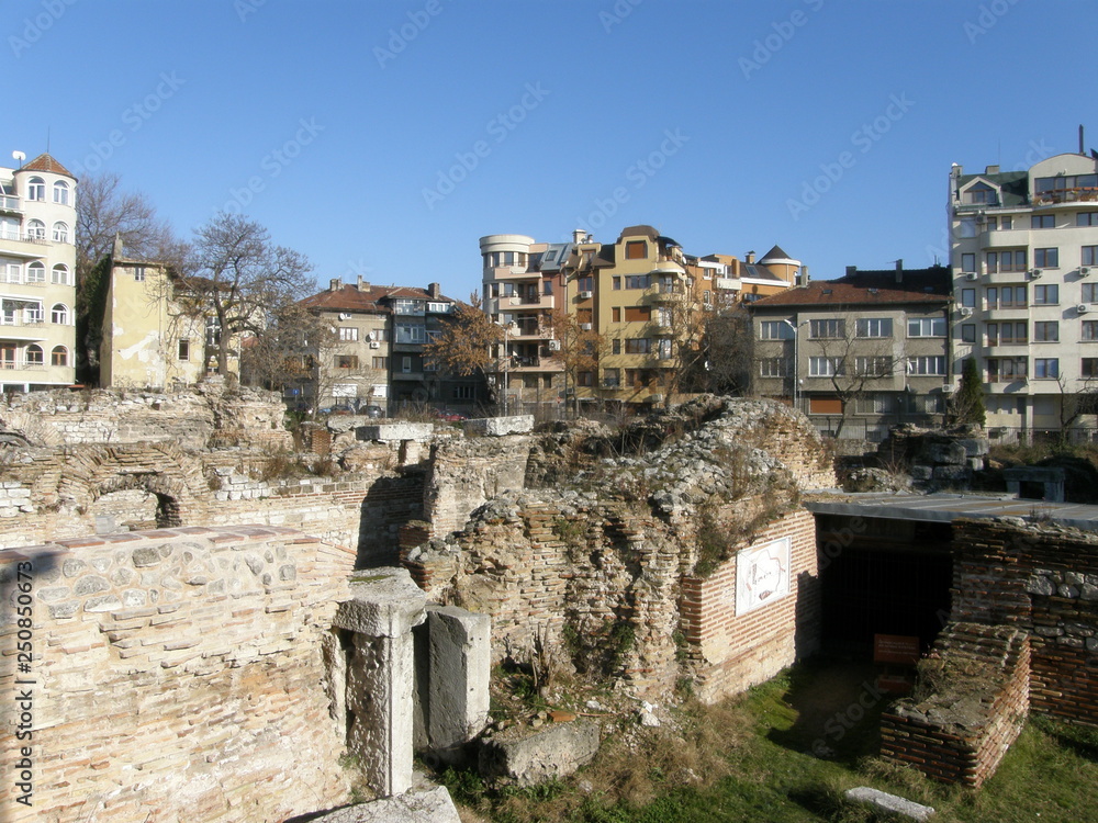 The ruins of an ancient stone structure below ground level against the background of modern multi-storey buildings on a Sunny day with a blue sky