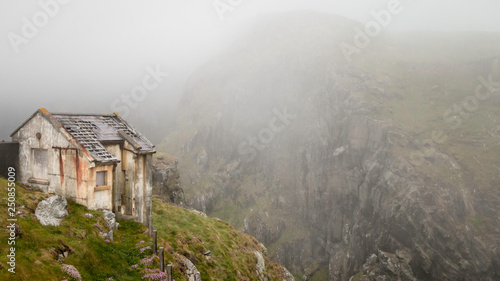 Old desolated shed on the edge of a ravine with misty cliffs in the background at Mizen Head in Ireland