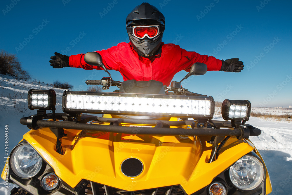 Photo of attractive young man in red warm winter clothes and black helmet with arms open on the ATV 4wd quad bike stand in heavy snow with deep wheel track. Moto winter sports.
