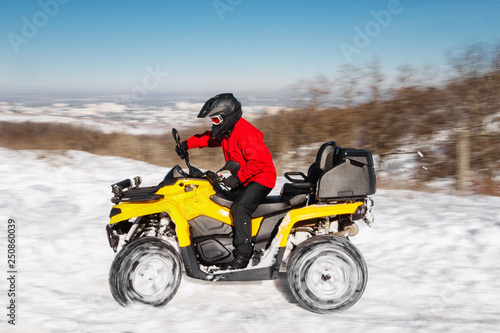 Photo of young extremal driver in red warm winter clothes and black helmet drive his ATV 4wd quad bike stand in heavy snow with deep wheel track. Extreme moto winter sports.