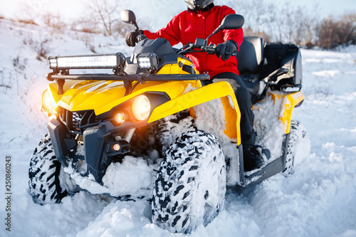 Close up photo of young man in red warm winter clothes and black helmet on the ATV 4wd quad bike stand in heavy snow with deep wheel track. Moto winter sports.