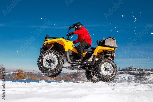 Photo of young extremal driver in red warm winter clothes and black helmet jump in the air with the ATV 4wd quad bike stand in heavy snow with deep wheel track. Extreme moto winter sports. photo