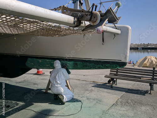 Worker wearing protection suit and mask is sanding down old paint from catamaran