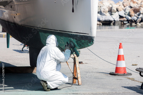 Worker wearing protection suit and mask is sanding down old paint from catamaran