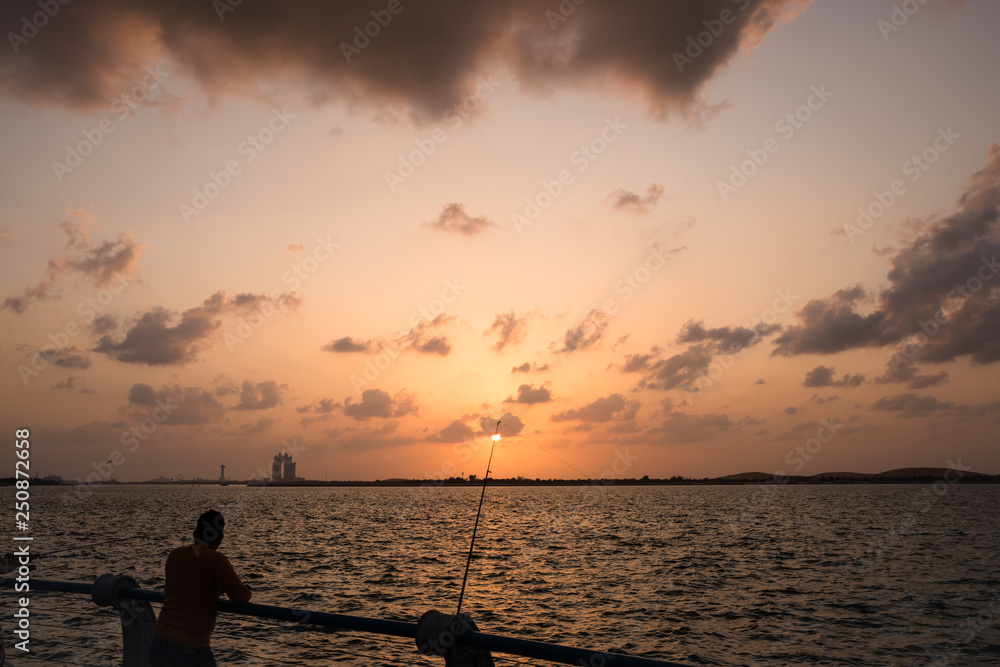 Fototapeta premium A man fishing while watching Sunset at Corniche beach during heavy dark clouds