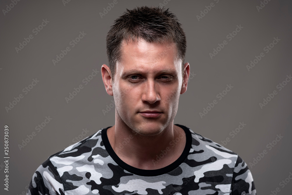 Fototapeta premium Dramatic moody close up portrait of angry young adult man with intense staring at camera on gray background. 