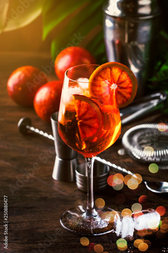 Aperol spritz cocktail in big wine glass with bloody oranges, summer Italian fresh alcohol cold drink. Wooden bar counter background with tools, summer mood concept with palm trees, selective focus