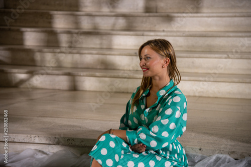 Smiling blonde girl sitting on the steps. Concept of fashionable dress. Woman in suit with 3/4 sleeves shirt and pleated polka dot skirt on aqua-green background.