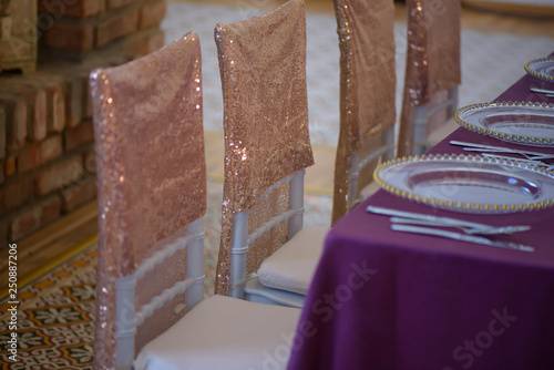Sitting arrangement at a fine dining restaurant or event featuring transparent plates with golden details, glassware and silverware in the order of use with purple table cloth and sequins chair covers