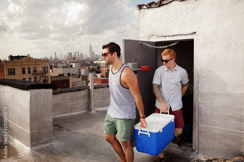 Two male friends carrying cooler out to city roof  photo