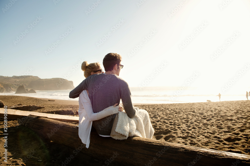 Young couple sitting on beach log and looking at view Young couple sitting on beach log and looking at view