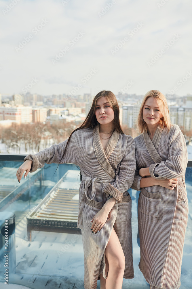 Cheerful young ladies in soft bathrobes resting at spa lounge, posing outdoor on the hotel terrace in the middle of winter snowy city