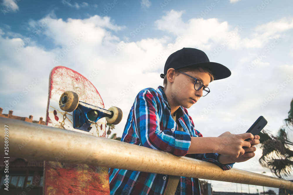 Handsome child with glasses and skater black hat resting on the railing  with cellphone, wearing a checkered shirt and gray shorts, close up his  skateboard Teen relaxing with phone enjoying outdoors Photos