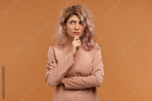 Body language. Thoughtful attractive fashionable hipster girl in stylish clothes looking upwards and touching her chin, having pensive deep in thoughts facial expression, coming up with idea