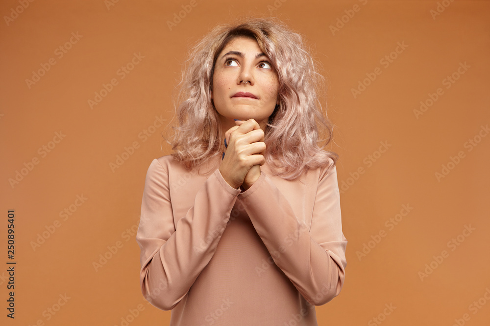 Isolated shot of beautiful fashionable young European female with nose ring and pinkish hair holding clasped hands and looking up with mournful hopeful facial expression, praying, worrying Isolated shot of beautiful fashionable young European female with nose ring and pinkish hair holding clasped hands and looking up with mournful hopeful facial expression, praying, worrying