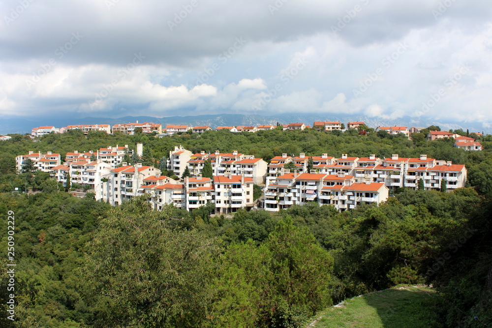 Multiple attached white apartment buildings on side of small hill surrounded with dense forest vegetation and family houses with cloudy blue sky in background on warm sunny day