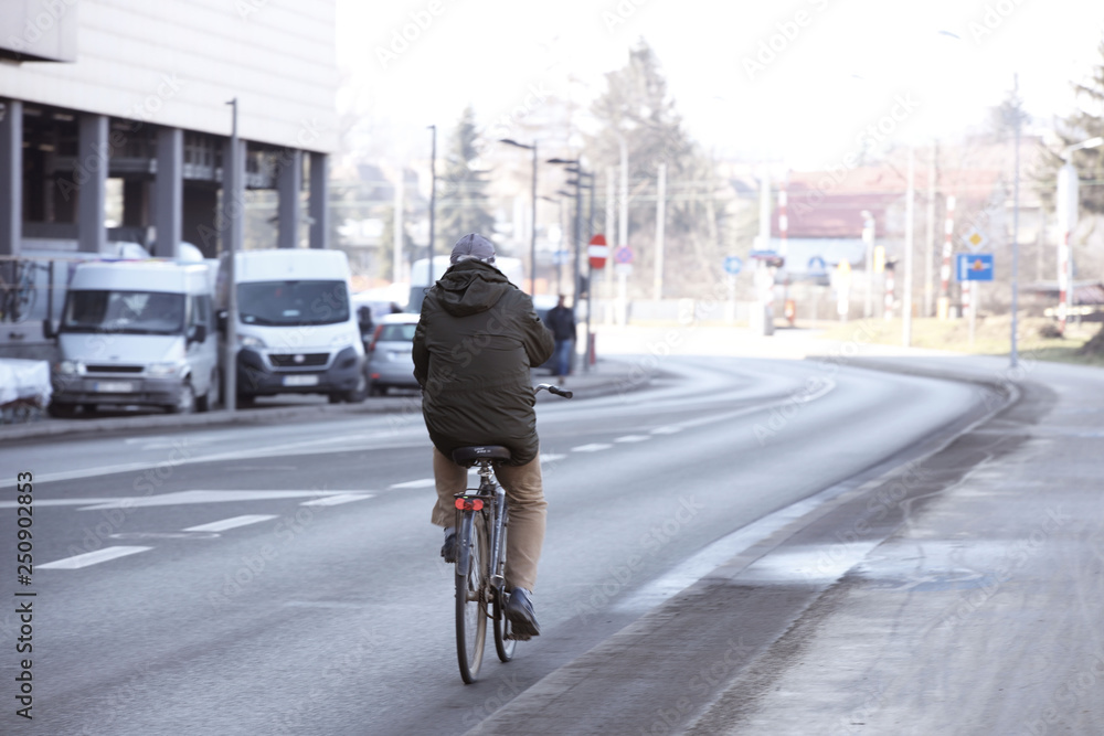 A man rides a bicycle along an asphalt city road near a shopping center with a truck parking. The worker gets to work in the morning at rush hour on environmental transport. Healthy lifestyle