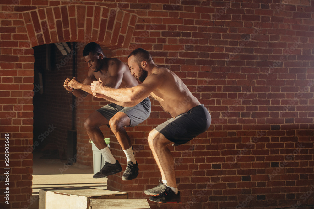 Obraz premium Two multi ethnic male sportsmen are jumping on plyo box, close up. African fitness instructor with his caucasian trainee doing box jump workout at cross fit gym