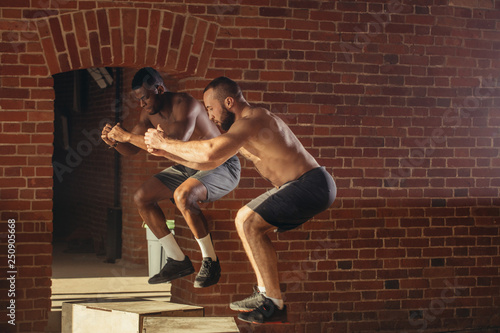 Two multi ethnic male sportsmen are jumping on plyo box, close up. African fitness instructor with his caucasian trainee doing box jump workout at cross fit gym