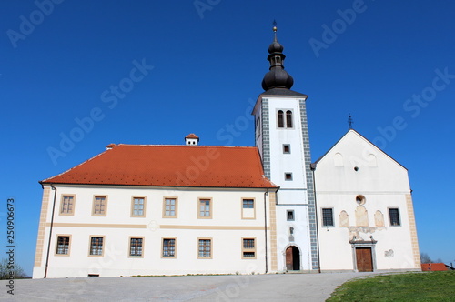 Old church with partially restored outer facade on top of small hill with large paved driveway surrounded with grass and clear blue sky in background on warm sunny day © hecos