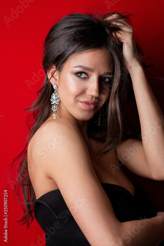Charming tanned model with long shiny hair wearing earrings posing on a red background