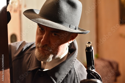 Closeup portrait of a mature caucasian  man in a fedora and raincoat (investigator, private detective or hitman) holding a gun. Interior scene selective focus