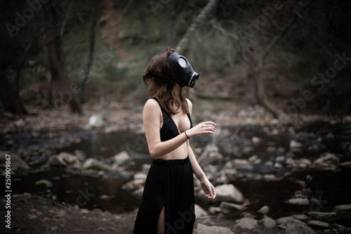Anonymous woman in black clothes and gas mask standing in amazing spooky forest