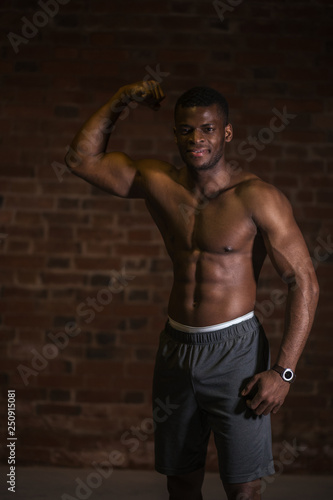 Handsome African American bodybuilder man, with naked muscular torso showing musculs against red brick wall.